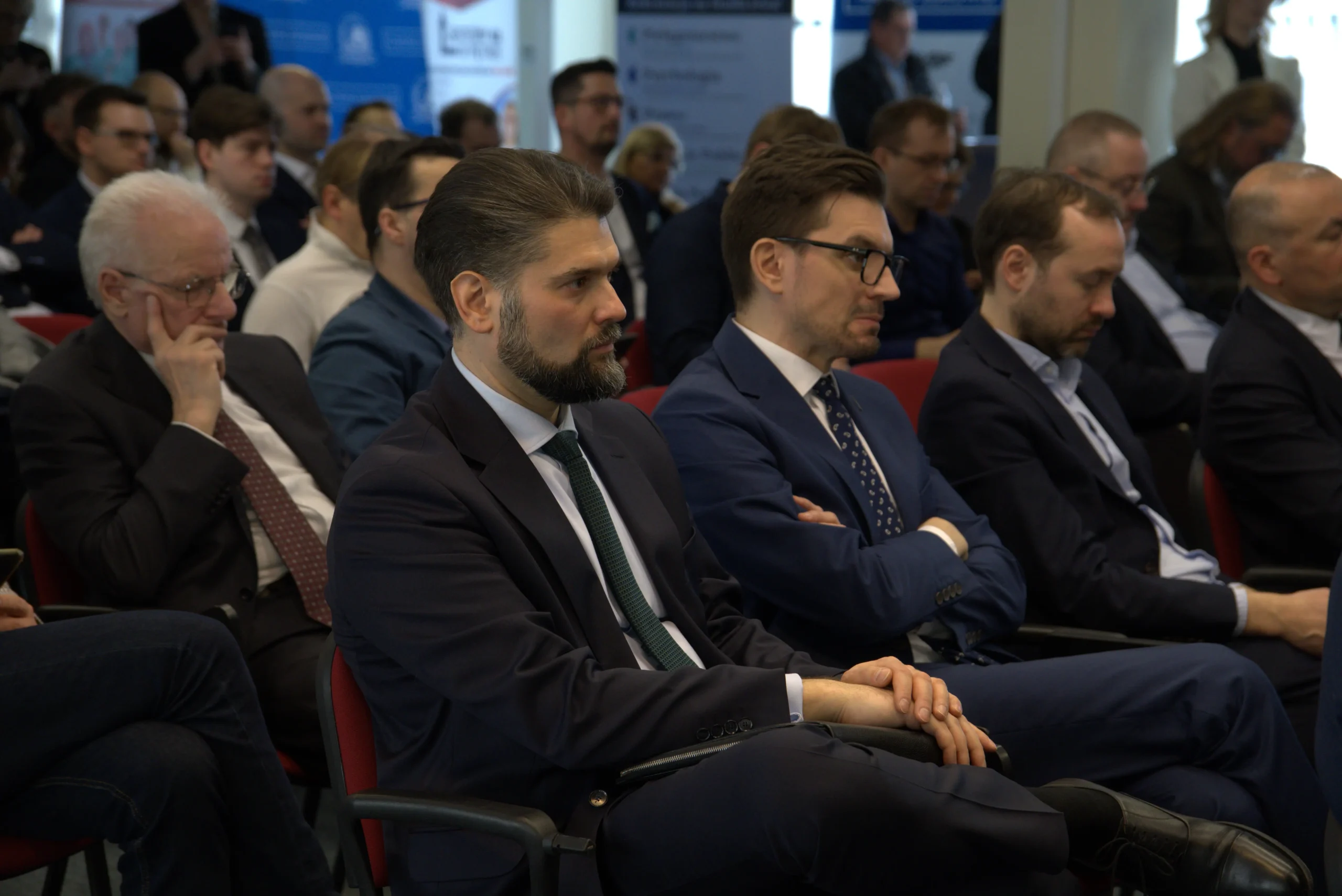 Uczestnicy konferencji biznesowej siedzący w rzędach, ubrani w garnitury, skupieni na słuchaniu prelegenta. Na pierwszym planie widać mężczyznę z brodą w ciemnym garniturze.