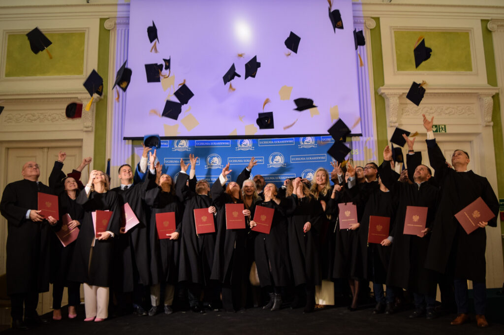 Grupa absolwentów i kadry akademickiej w czarnych togach rzuca birety w górę podczas uroczystości ukończenia studiów na Uczelni Łukaszewski