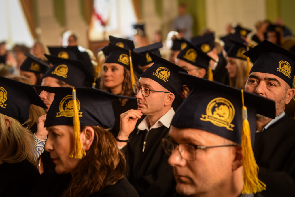 Grupa absolwentów w czarnych togach i biretach ze złotymi chwostami siedzi w rzędach, uważnie słuchając przemówienia podczas uroczystości.