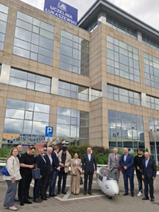 Grupa uczestników Dnia Otwartego pozuje do zdjęcia przed budynkiem Uczelni Łukaszewski, obok niewielkiego pojazdu aerodynamicznego.