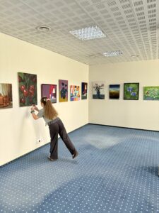 Ekspozycja prac malarskich absolwentów Uczelni Łukaszewski oraz osoba fotografująca jeden z obrazów.